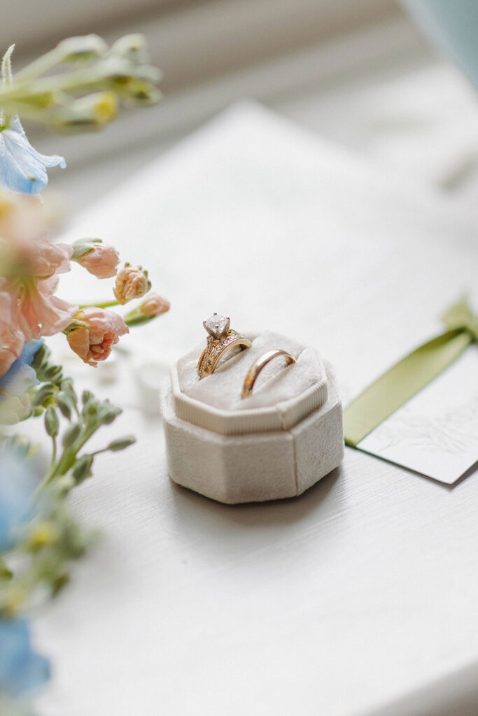 Wedding rings displayed in a soft velvet ring box beside pastel flowers and handwritten vows.