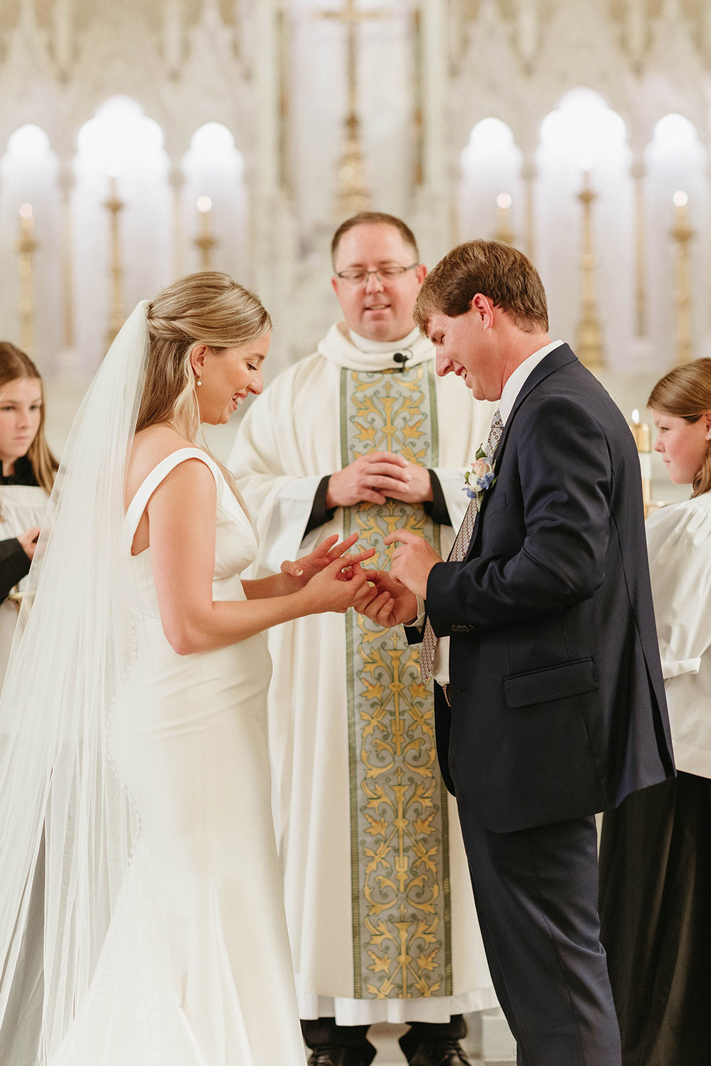 Bride and groom exchange rings during a church wedding ceremony, standing at the altar with a priest behind them.