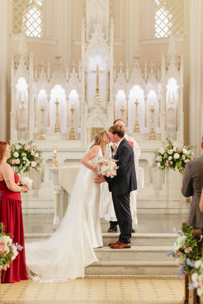 Bride and groom share their first kiss at the altar inside a bright church, surrounded by floral arrangements.