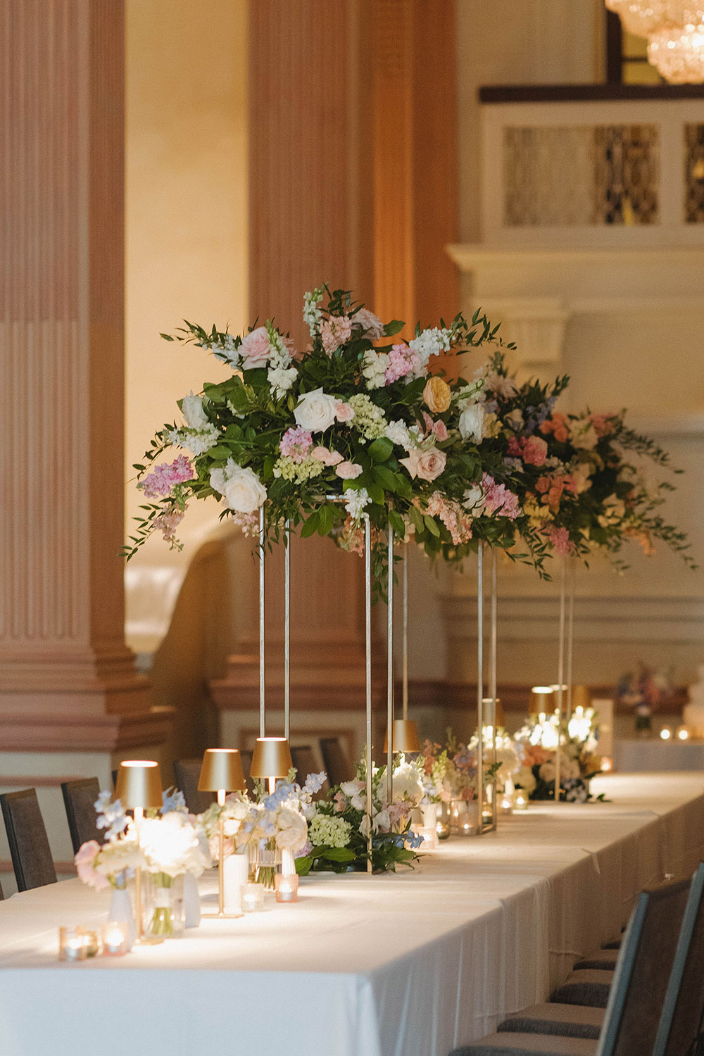 Elegant wedding reception tables with tall floral centerpieces, soft candlelight, and pastel arrangements inside a grand ballroom.