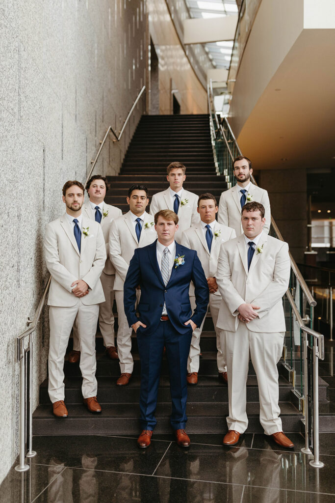 Groom stands with his groomsmen on a modern staircase, wearing a navy suit while the groomsmen wear light suits with blue ties.