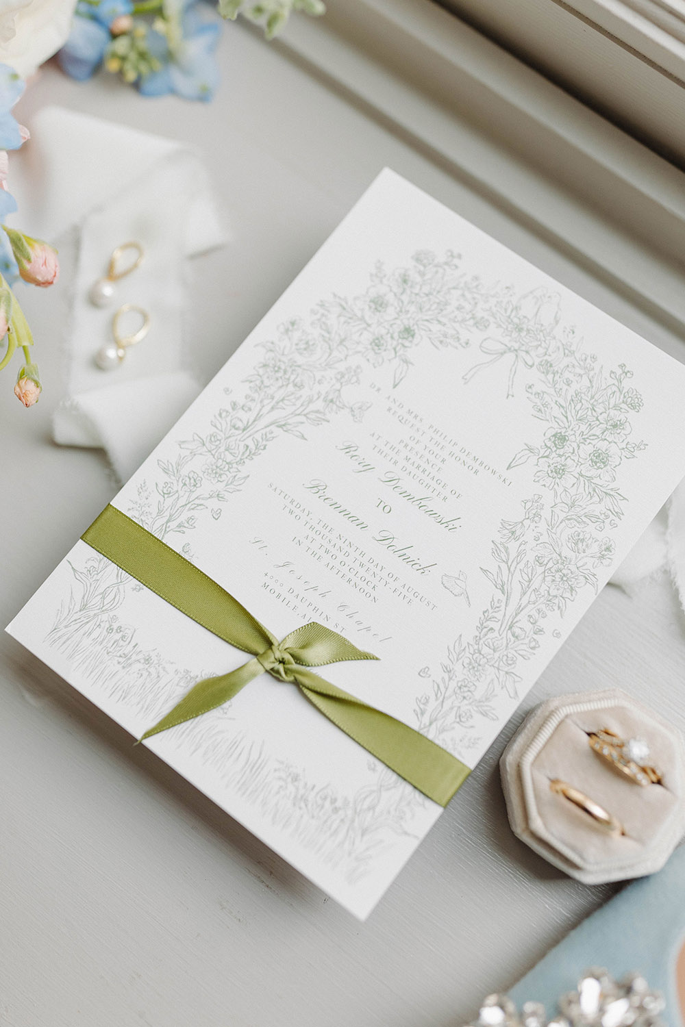 Elegant wedding invitation with delicate floral illustration and green ribbon, styled with rings, pearl earrings, and pastel flowers on a windowsill.