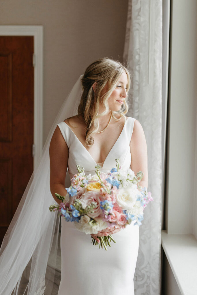 Bride in a minimalist white wedding gown holds a pastel garden bouquet while standing by a window.
