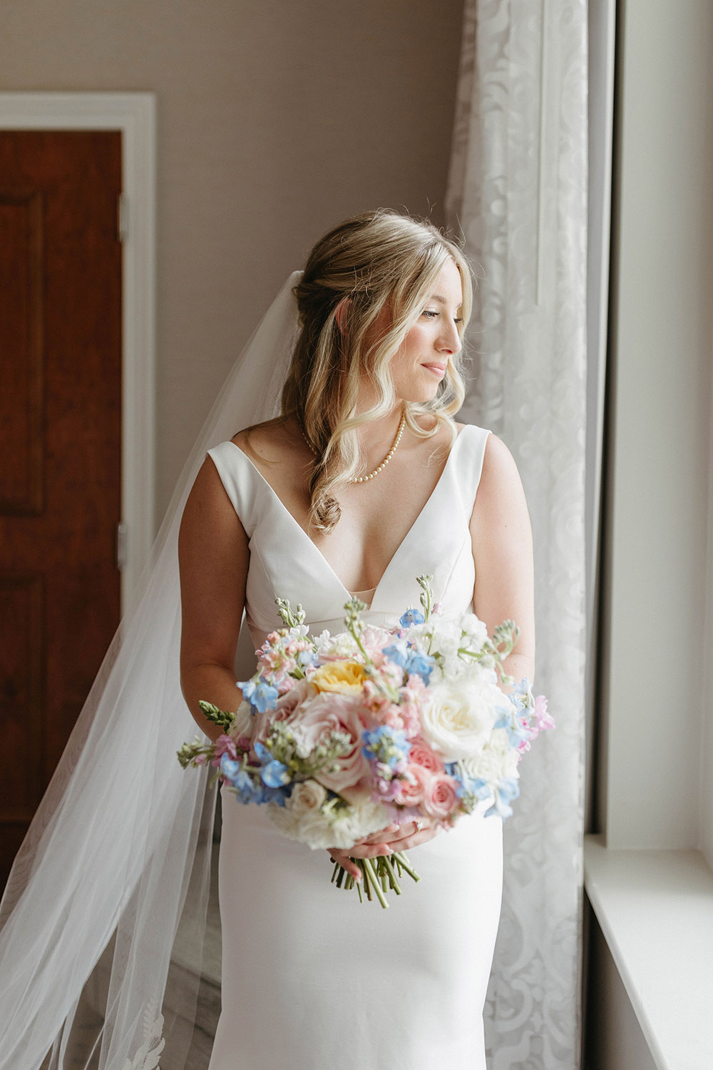 Bride in a minimalist white wedding gown holds a pastel garden bouquet while standing by a window.
