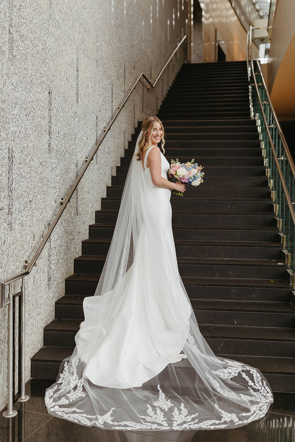 Bride in a sleek white wedding gown with a long lace-trimmed veil stands on a modern staircase holding a pastel bouquet.