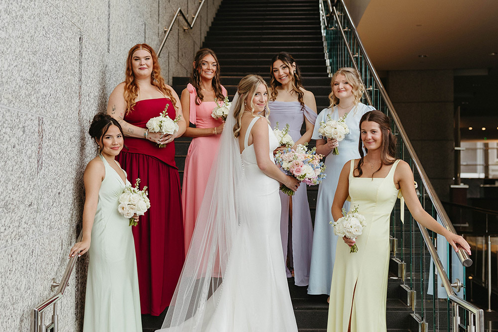 Bride stands on a staircase with her bridesmaids in pastel dresses, each holding white bouquets, inside a modern venue.