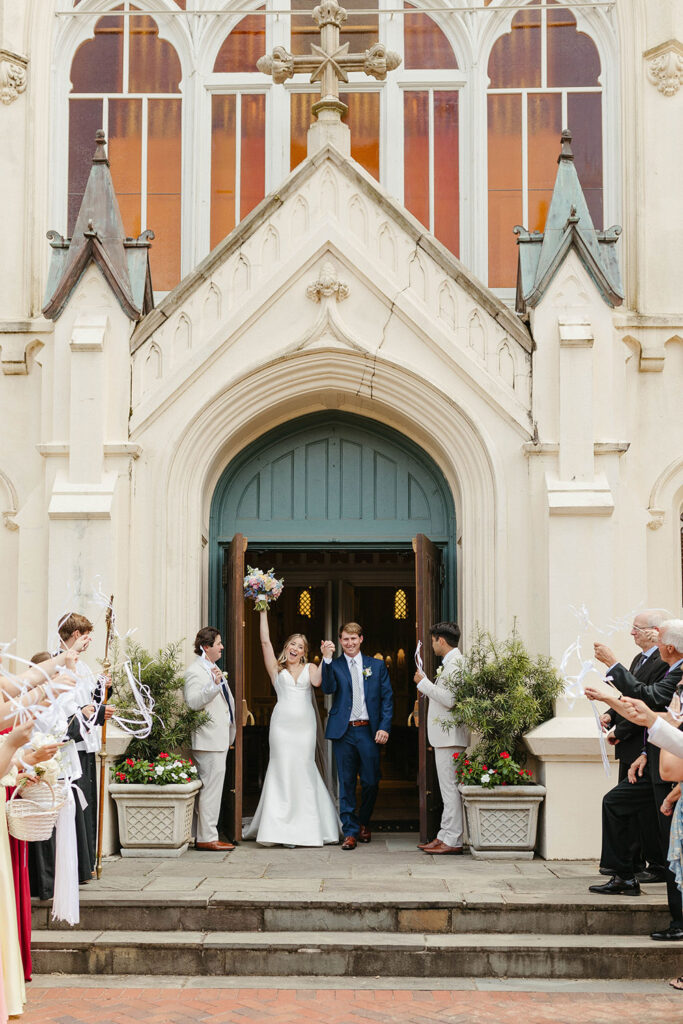 Newlyweds exit a church together, cheering guests tossing ribbon wands as they celebrate outside the entrance.