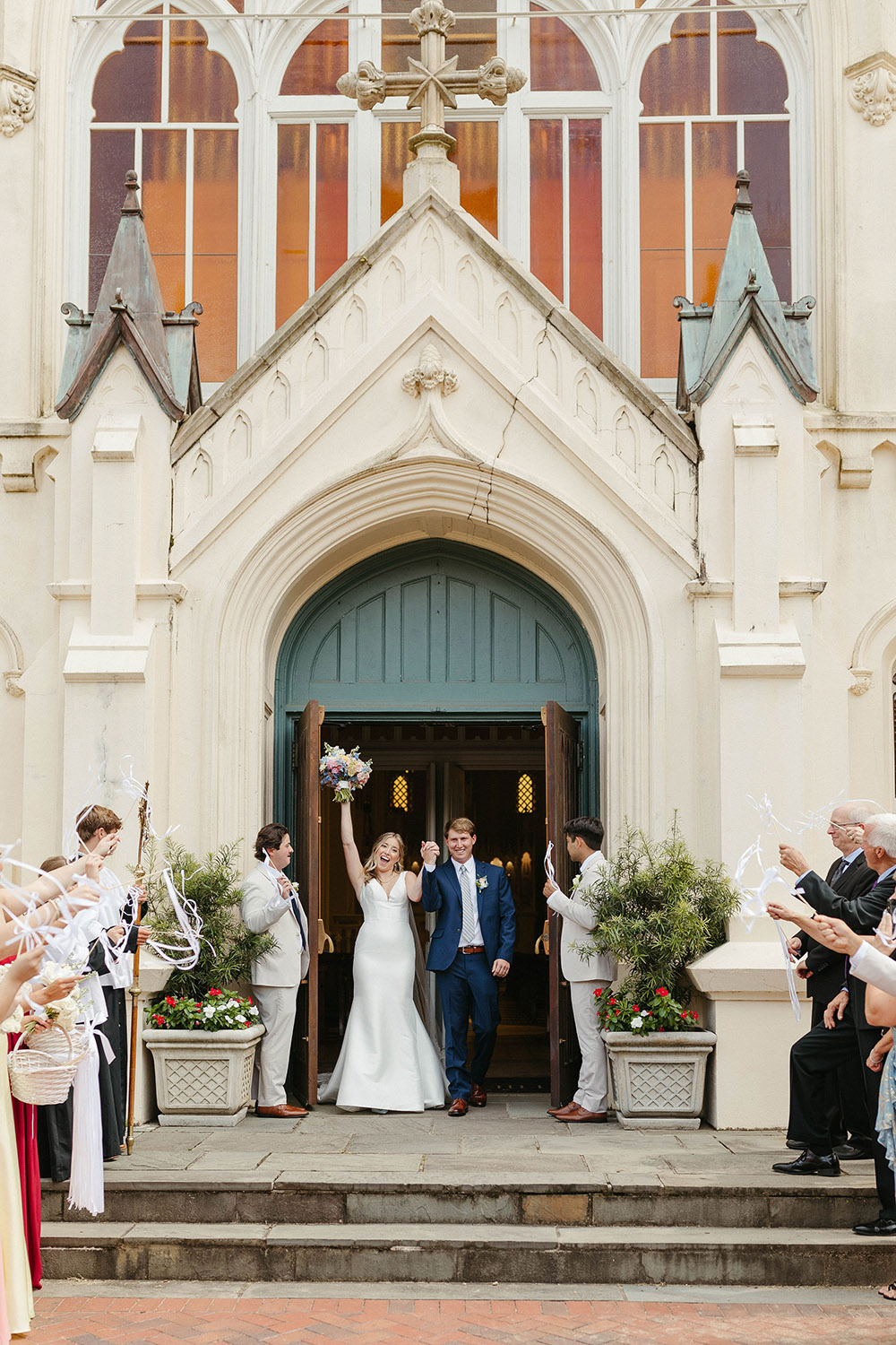 Newlyweds exit a church together, cheering guests tossing ribbon wands as they celebrate outside the entrance.