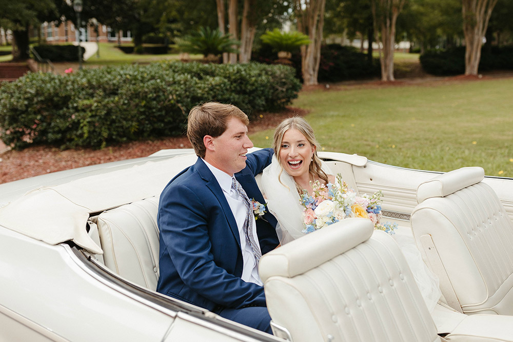 Newlyweds laugh together while riding in a white vintage convertible, the bride holding a colorful spring bouquet.