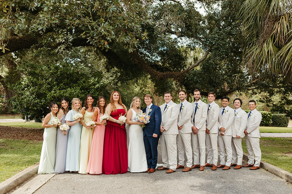 Bride and groom pose with their wedding party outdoors, bridesmaids in pastel dresses and groomsmen in light suits beneath oak trees.