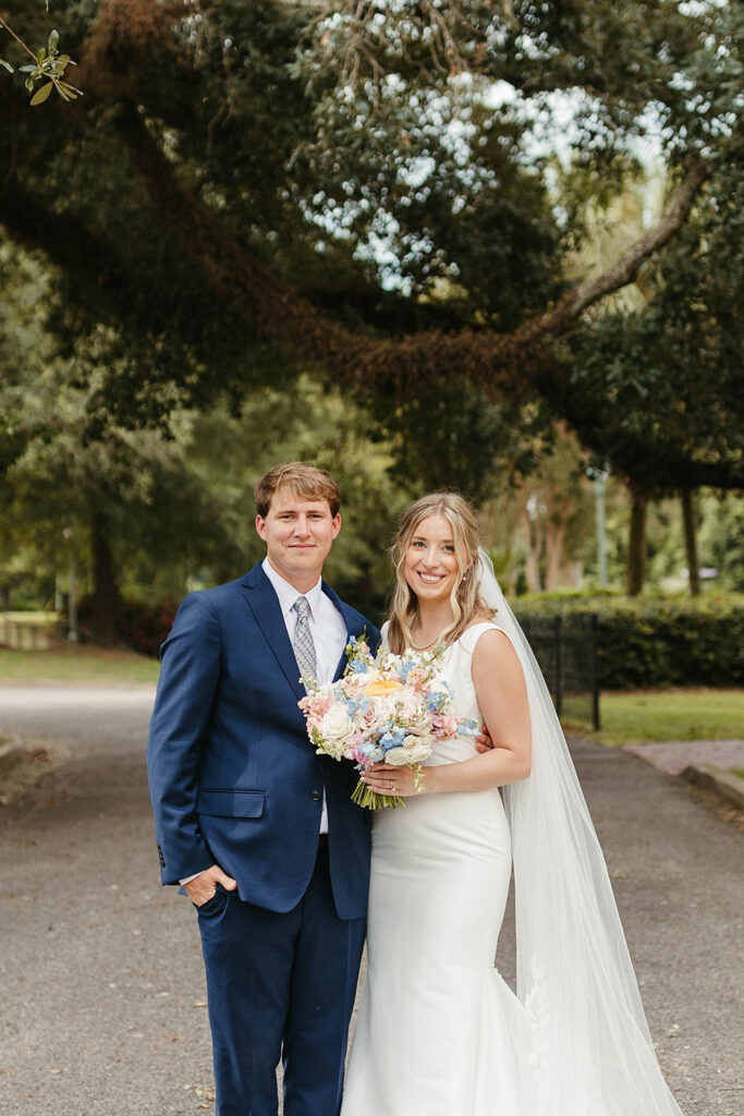 Bride and groom standing together outdoors beneath oak trees, the bride holding a pastel wedding bouquet and wearing a long veil.