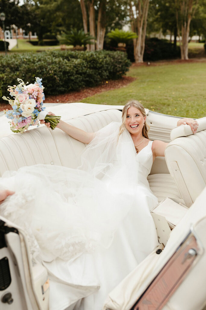Bride reclines in a white vintage convertible, smiling and holding her pastel wedding bouquet outdoors.