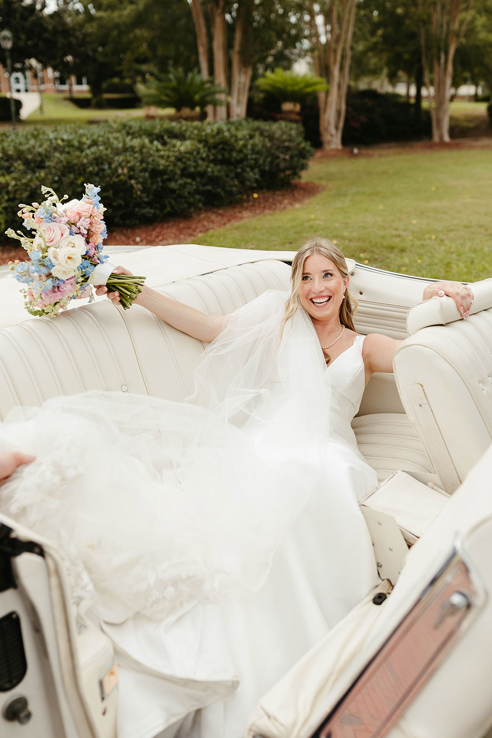 Bride reclines in a white vintage convertible, smiling and holding her pastel wedding bouquet outdoors.