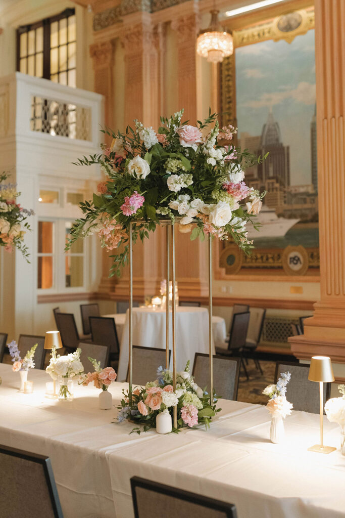 Tall floral wedding centerpiece with pastel blooms and greenery displayed on a long reception table inside an elegant ballroom.
