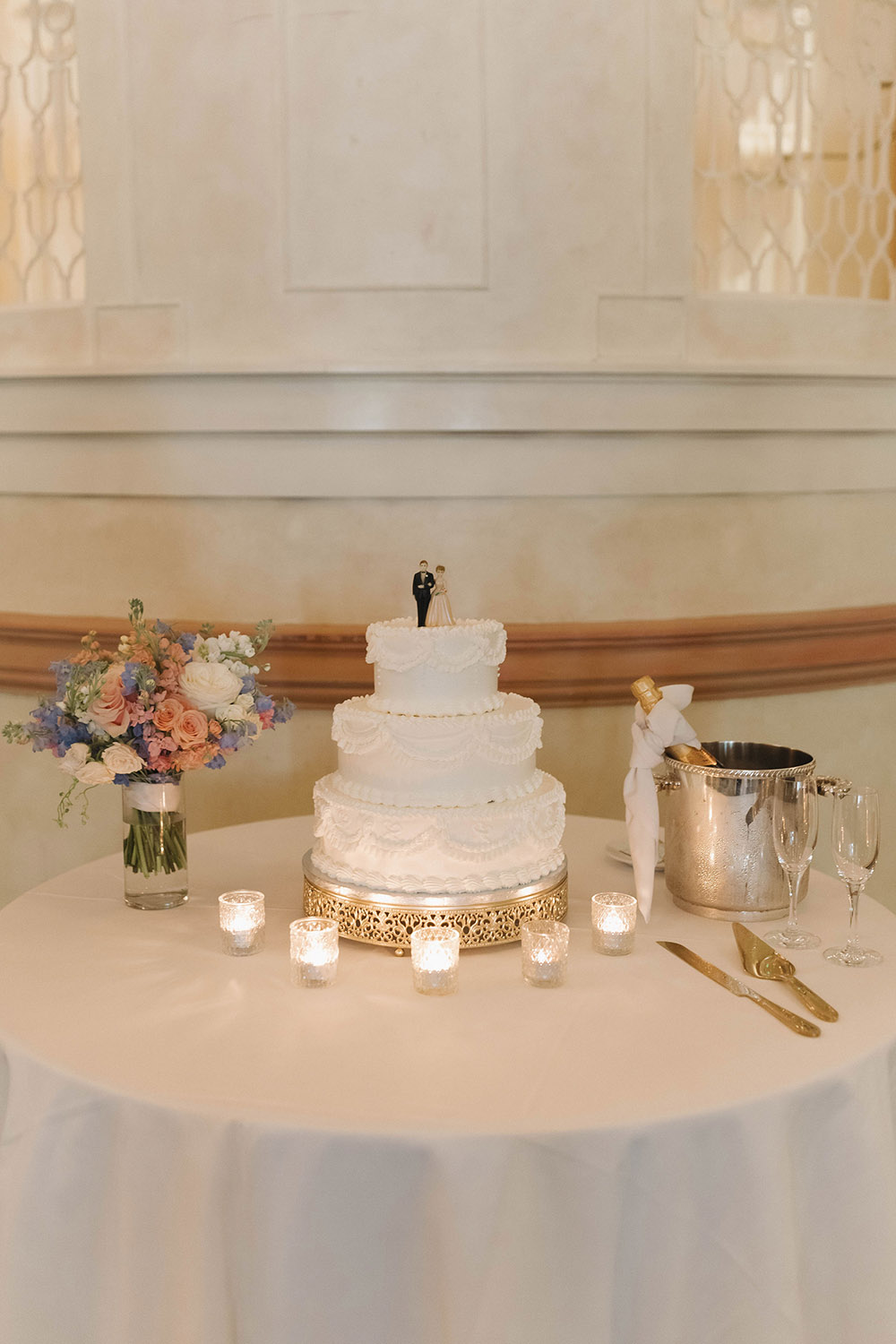Three-tier white wedding cake with classic piping displayed on a round table with candles, floral accents, and champagne glasses.