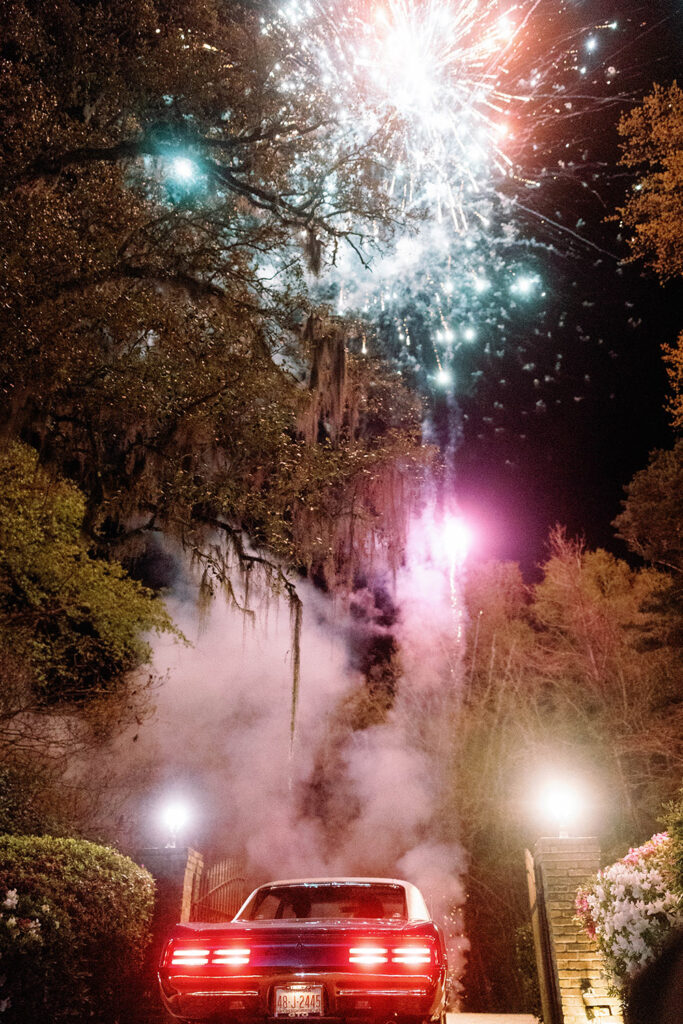 A classic blue car drives away under a sky full of bright fireworks, creating a dramatic and celebratory nighttime wedding exit scene with smoke and glowing lights along the driveway.