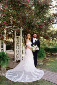 Bride and groom stand smiling beneath a garden arbor with lush flowering trees overhead. The bride wears an off-the-shoulder lace wedding gown with a long, floral-embroidered train and holds a white and blush bouquet. The groom stands beside her in a dark suit with a boutonniere, surrounded by greenery and ferns in a serene outdoor setting.
