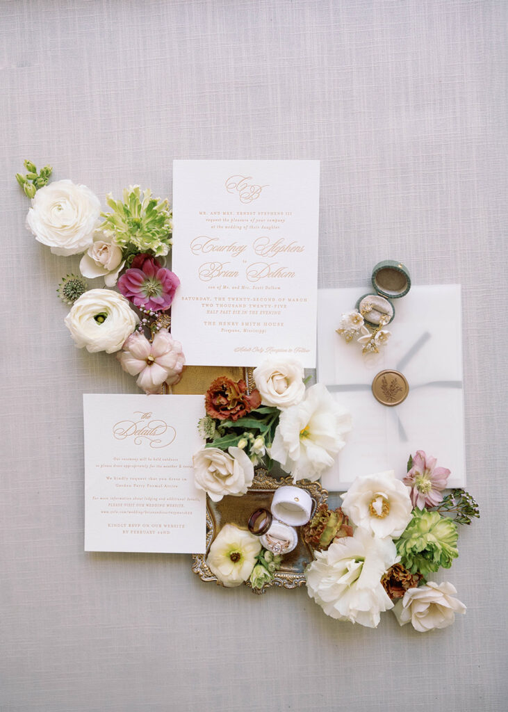 Elegant wedding invitation suite flatlay featuring cream and amber stationery, wedding rings in velvet boxes, and assorted pastel florals arranged on a light linen backdrop.