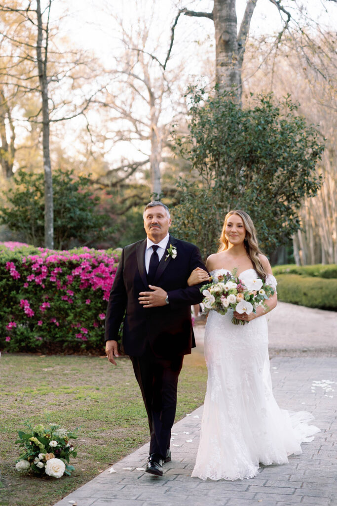 Bride walking down the aisle outdoors with her father, holding a floral bouquet, surrounded by blooming garden scenery during a sunset wedding ceremony.