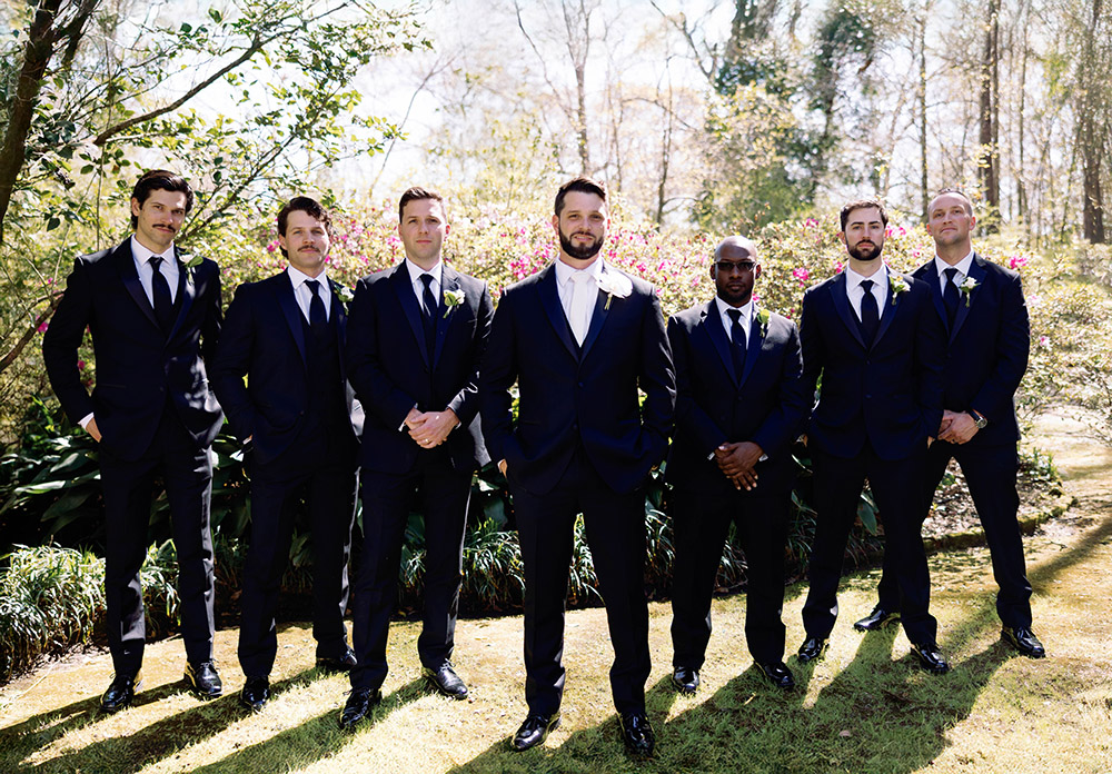Groom and groomsmen dressed in black tuxedos posing outdoors in a garden setting, standing confidently in sunlight with blooming greenery behind them.