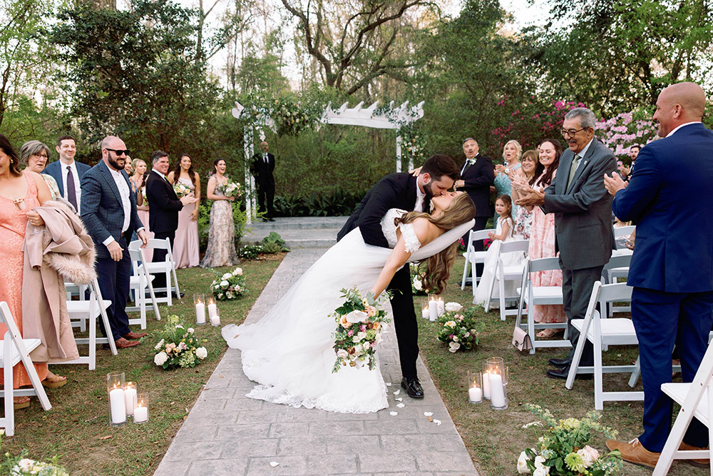 Groom dips and kisses the bride during their outdoor wedding ceremony recessional, surrounded by cheering guests, floral aisle arrangements, and candle decor in a lush garden setting.
