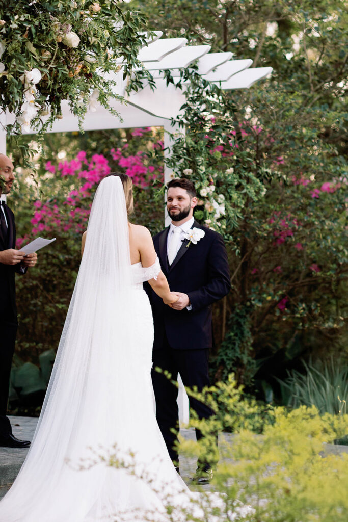 Bride and groom share their first kiss beneath a white arbor adorned with cascading greenery and flowers during their outdoor wedding ceremony.
