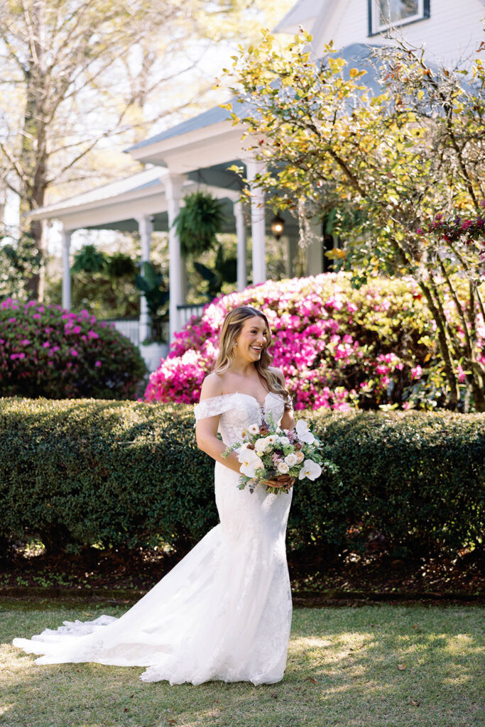 Bride in an off-the-shoulder lace wedding gown smiling and holding a pastel floral bouquet, standing outdoors in a garden with blooming pink azaleas and a Southern-style white porch in the background.