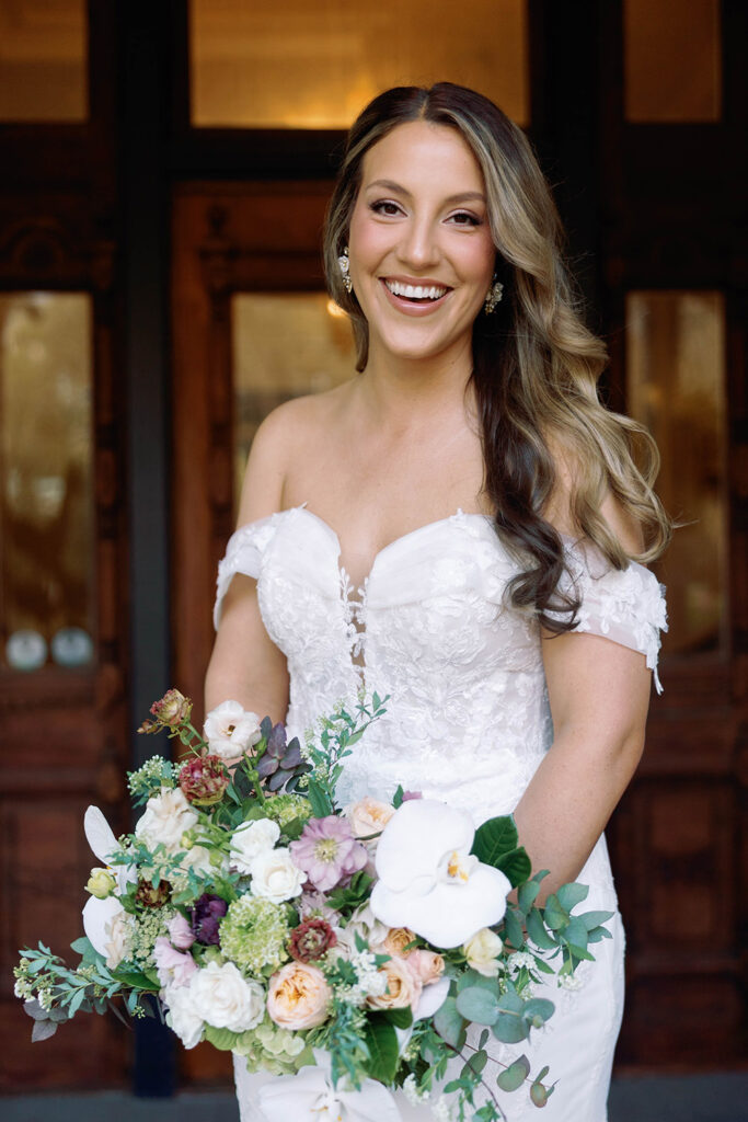 Bride smiling in an off-the-shoulder lace wedding gown, holding a lush bouquet of white orchids and pastel flowers, posing outdoors in natural light.