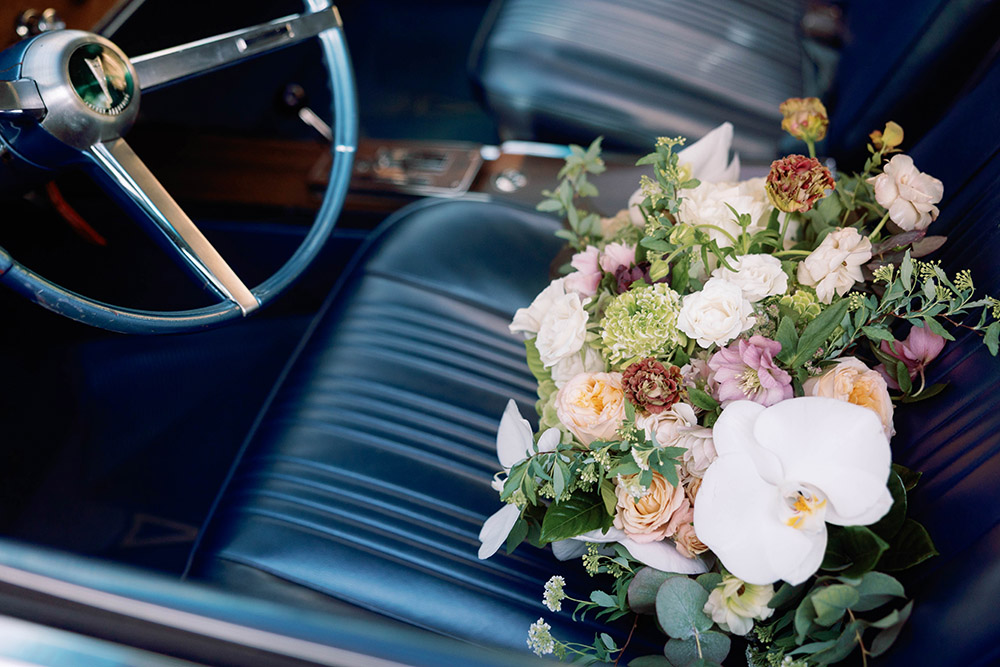 Bridal bouquet featuring white orchids, blush and cream roses, and greenery resting on the blue leather seat of a vintage car with a classic steering wheel visible.
