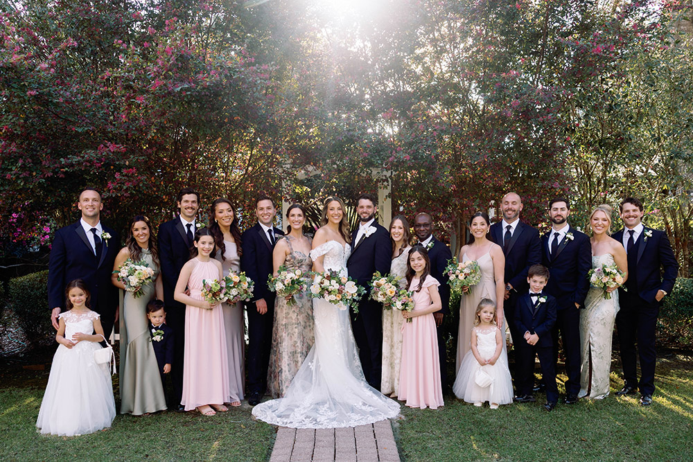 “Large wedding party portrait with the bride and groom surrounded by bridesmaids in neutral and blush dresses and groomsmen in navy suits, posed outdoors under sunlit trees holding bouquets.”