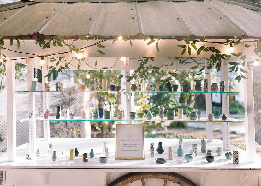 Display of handcrafted ceramic wedding favors arranged on glass shelves of a white cart decorated with greenery and string lights at an outdoor reception.
