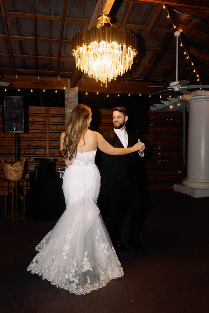 Bride and groom share their first dance under a glowing chandelier, smiling at each other as she twirls in her lace mermaid wedding gown during the evening reception.