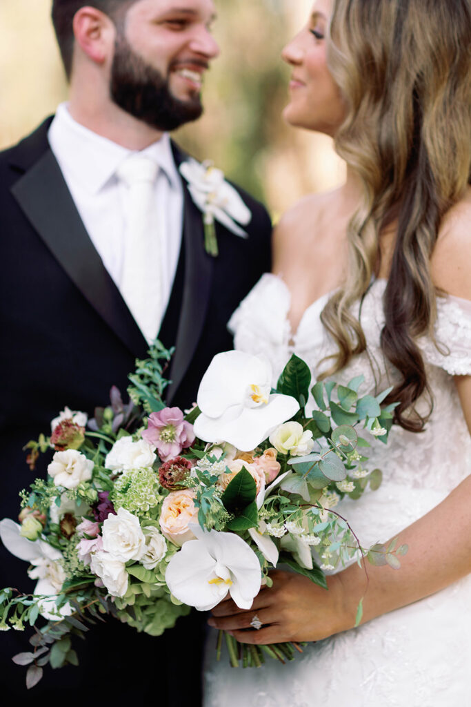 Close-up of a bride and groom smiling at each other while the bride holds a lush bouquet of white orchids, roses, and greenery, with the groom in a black tuxedo and the bride in an off-the-shoulder lace gown.