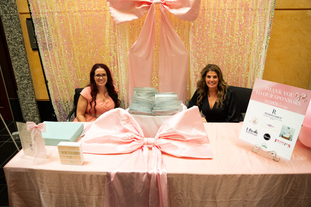 Two women seated behind a pink-draped welcome table with a large bow backdrop, gift bags, and a “Thank You to Our Sponsors” sign at a bridal event.