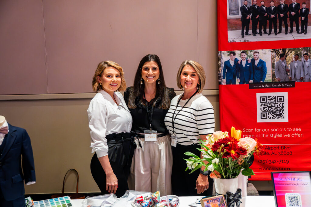 Three women smiling together at a bridal show vendor booth, standing beside a red tuxedo and suit rental display with floral arrangements and promotional materials.
