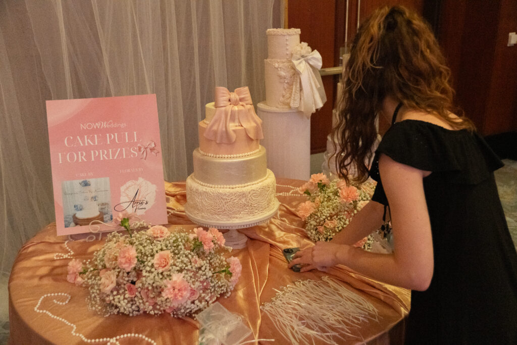 Attendee selects a ribbon prize from a blush pink cake pull display featuring tiered cakes, floral arrangements, and a “Cake Pull for Prizes” sign at a wedding event.