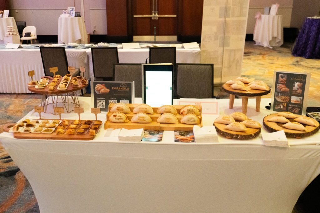 Empanada catering display at a bridal show or wedding expo, featuring assorted savory empanadas arranged on tiered wooden stands atop a white linen table in a ballroom setting.