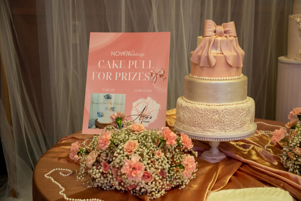 Blush pink three-tier display cake with a large fondant bow, set on a satin-draped table with pink floral arrangements and a NOW Weddings “Cake Pull for Prizes” sign.
