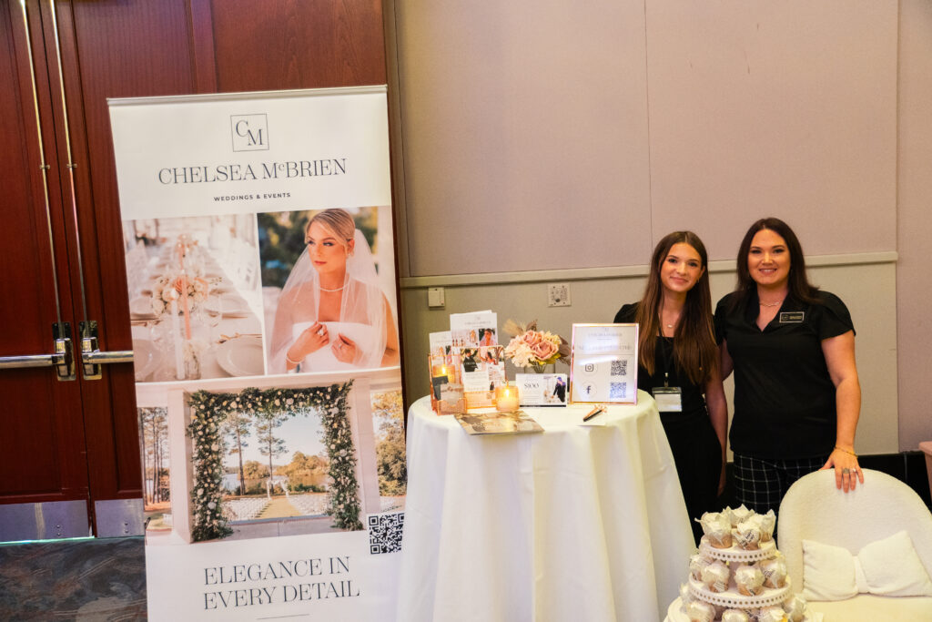 Wedding planning vendor booth at a bridal show, featuring two planners standing beside a branded banner and a styled cocktail table with brochures, florals, candles, and display signage.