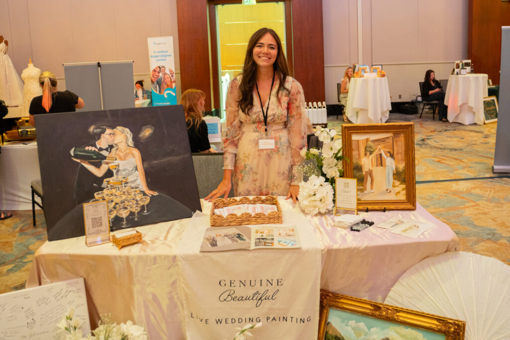 Wedding artist standing behind a bridal show booth featuring live wedding paintings, framed artwork, floral accents, brochures, and display signage.