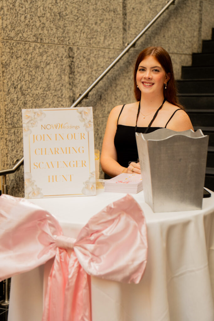 NOW Weddings team member smiles at a welcome table featuring a scavenger hunt sign, blush pink bow décor, and printed materials at a bridal event.