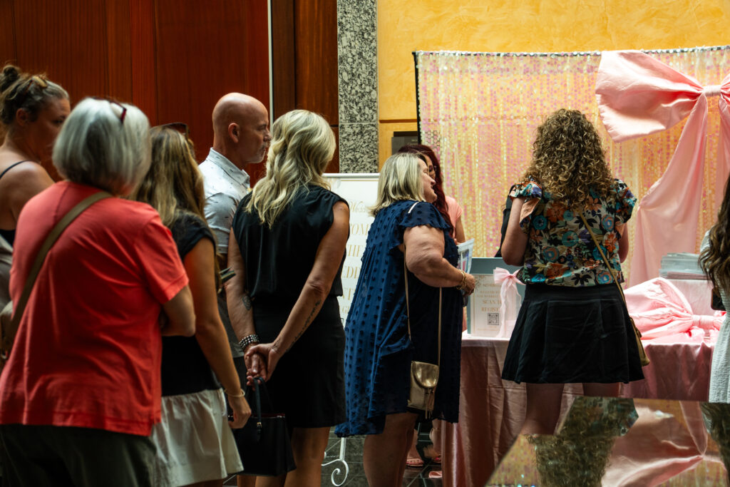 Wedding show attendees standing in line at a decorated booth with pink backdrop, large bow, and informational signage.
