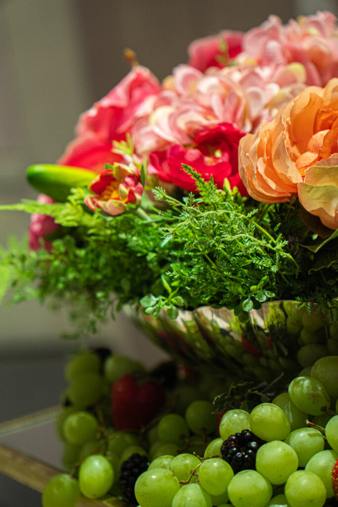 Close-up of a colorful floral arrangement with pink and peach blooms, greenery, and fresh grapes displayed below.