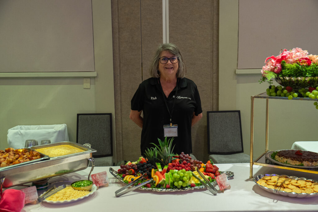 Caterer standing behind a dessert and fruit display with assorted cheeses, crackers, and plated dishes at an event table.