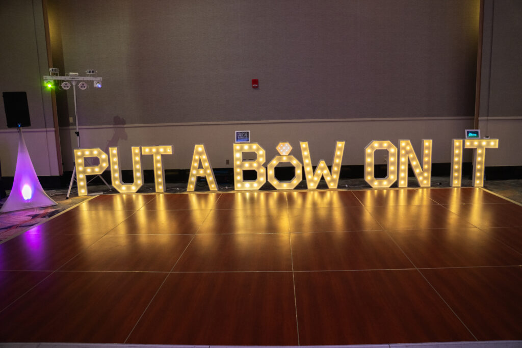 Illuminated marquee letters spelling “Put a Bow on It” displayed across a ballroom dance floor at a bridal event.