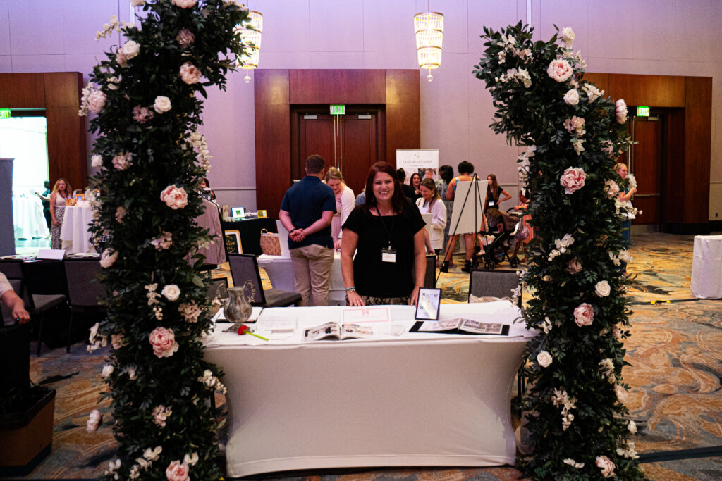 Wedding vendor booth framed by lush floral columns with blush and white blooms, featuring a designer display table at a bridal show or wedding expo.