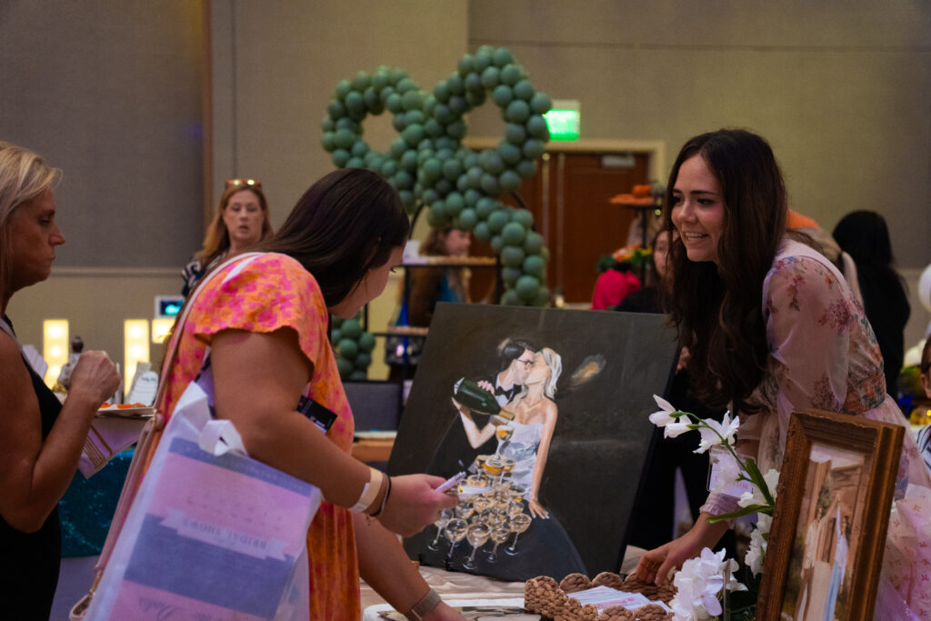 Wedding vendor showcases a painted wedding portrait at a bridal show booth as an attendee looks on, with floral accents and balloon décor in the background.