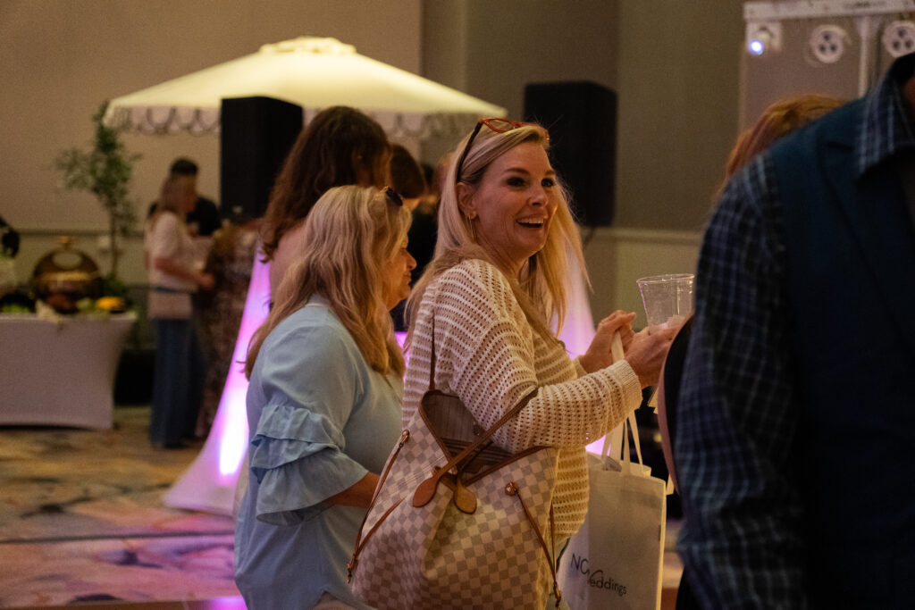 Wedding expo guests chatting and smiling while holding drinks and a NOW Weddings tote bag in a softly lit event space.
