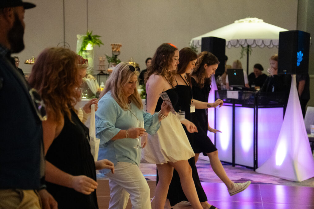 Guests line dance together on the dance floor at an event, moving in unison with DJ booth and purple uplighting in the background.