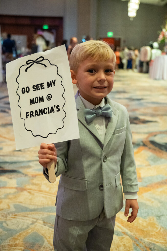 Young boy in a light gray suit and bow tie holding a sign that reads “Go see my mom @ Francia’s” at a wedding expo.